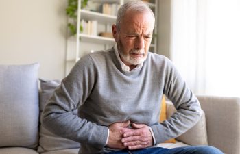 Senior man experiencing stomach or abdominal pain while sitting on his sofa, health problems in old age