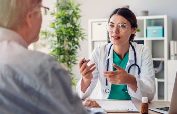A doctor wearing a white coat and stethoscope talks to a patient across a desk in a medical office. A pill bottle and papers are on the desk.