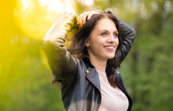 Beautiful, powerful middle-age woman in black leather jacket smiling broadly, arms above head. Springtime, outdoors