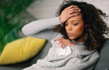 A woman sits on a couch, holding a tissue and touching her forehead, appearing unwell or fatigued.
