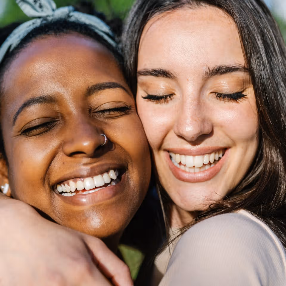 Two women with closed eyes smile and hug each other outdoors in natural light.