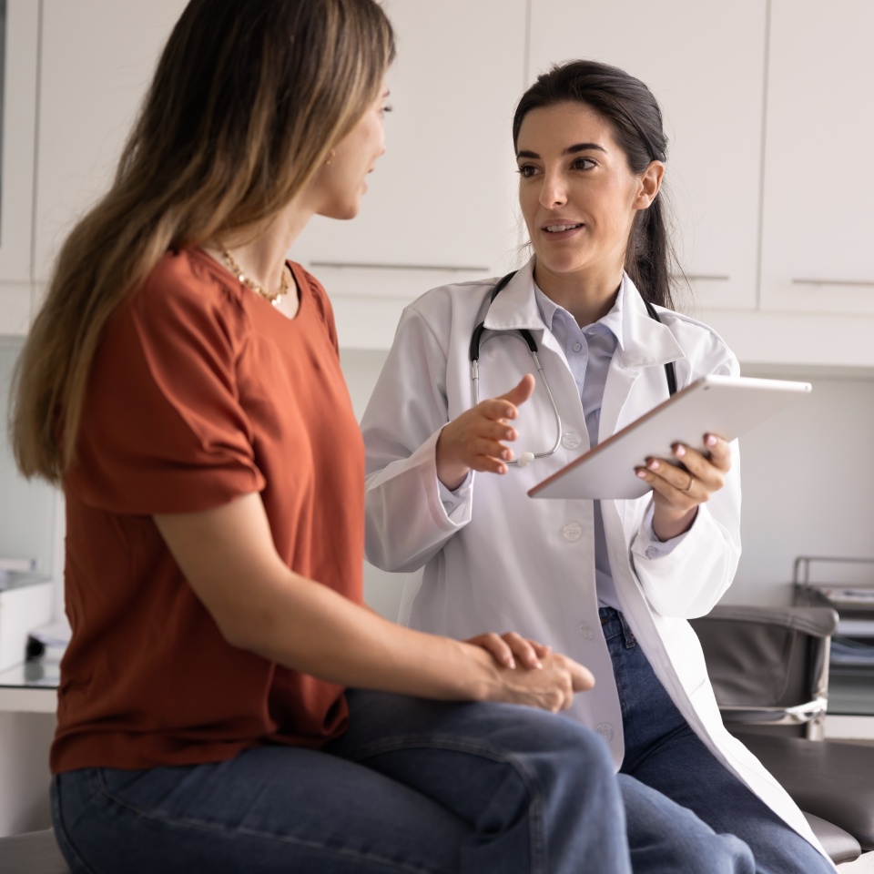 Professional Latina therapist holding modern digital tablet during conversation with woman patient, accessing electronic health record system, explain treatment plan or health test results. Healthcare
