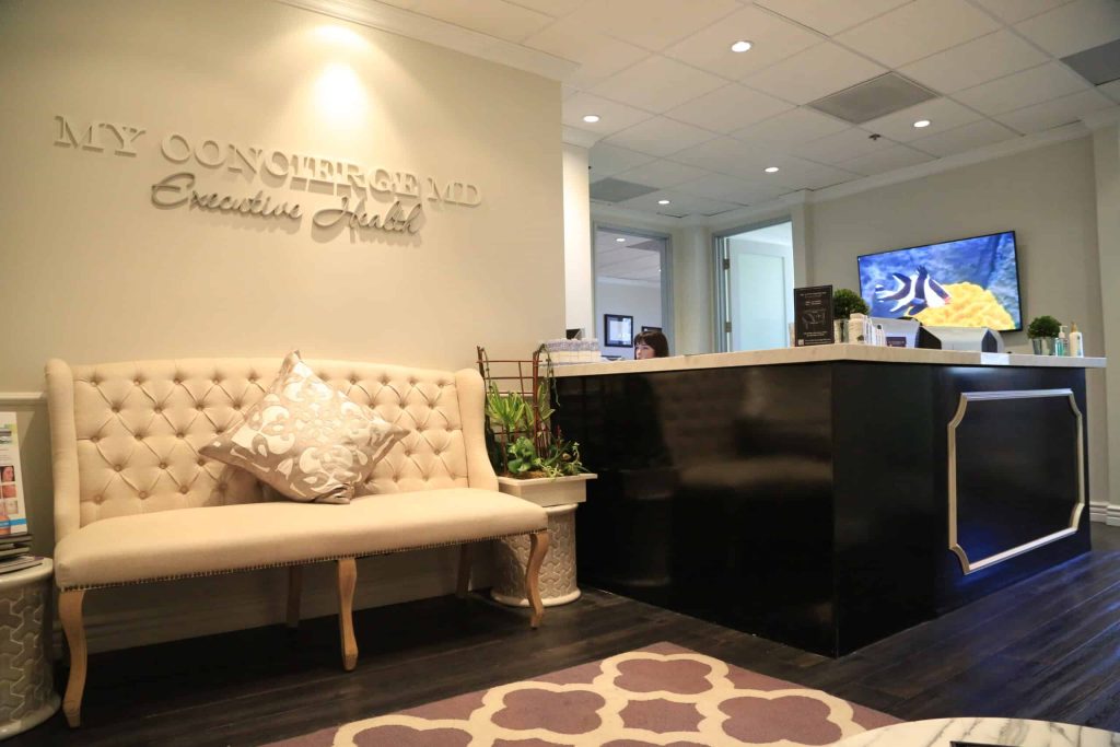 Reception area of a medical office with a beige tufted bench, black front desk, TV on the wall, and decorative plants.