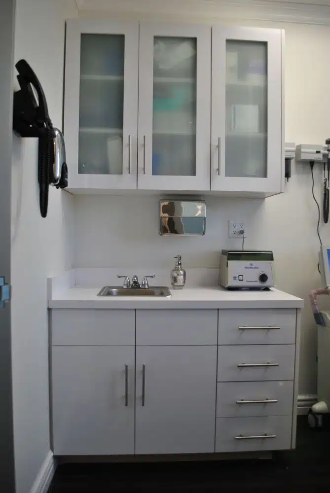 A clean, white medical exam room countertop with a sink, soap dispenser, drawers, cabinet, and medical equipment mounted on the wall.