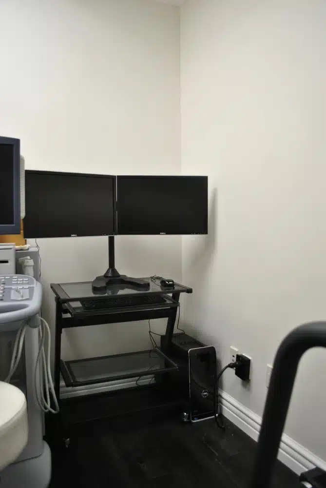 A small office setup with two black monitors on a stand, a keyboard, and a computer tower on a cart against a plain white wall.