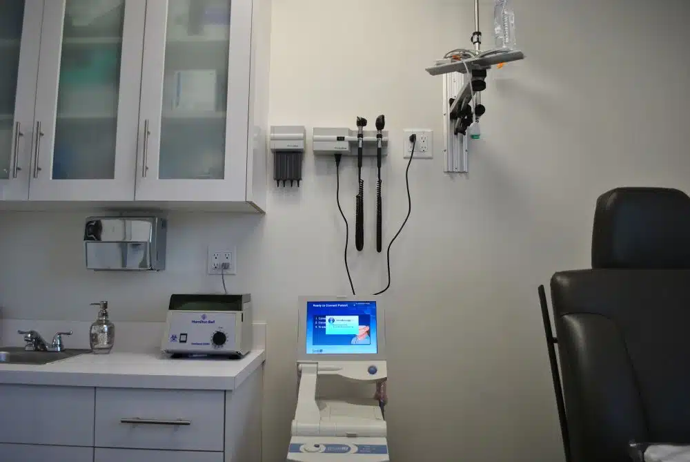 A medical examination room with a counter, sink, equipment, monitor, exam chair, and diagnostic tools mounted on the wall.