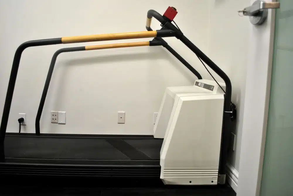 A treadmill with unusual large orange handlebars and a bulky white CRT monitor attached beside it, set against a white wall in a room.