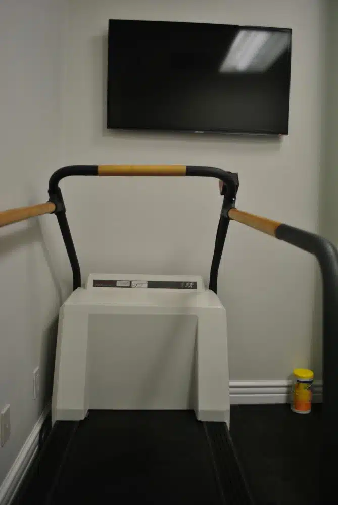 A treadmill facing a wall-mounted flat-screen TV, with a container placed on the floor in the corner of the room.