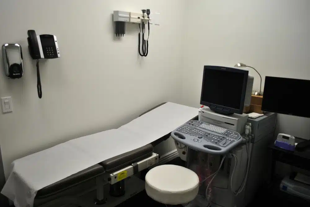 A medical examination room with an exam table, a wall-mounted blood pressure monitor, a computer, and ultrasound equipment.