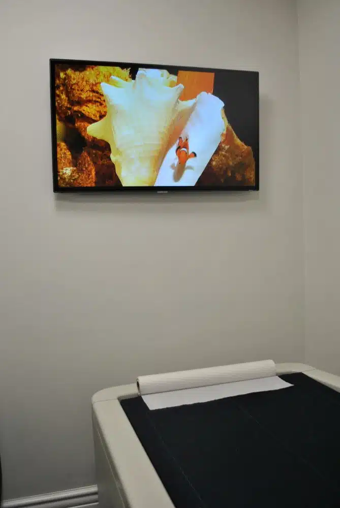 A TV mounted on a light-colored wall displays an image of a clownfish and a seashell above a medical examination table with a paper cover.