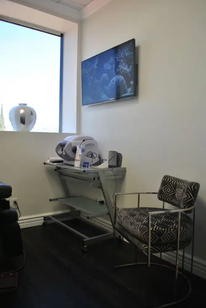 A small room with a patterned chair, a glass desk holding medical equipment, a wall-mounted TV, and a window with a white vase on the sill.