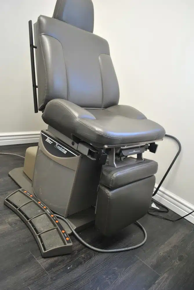 A gray, adjustable medical examination chair is positioned in a room with dark wood flooring and white walls. Various foot pedals and an electrical cord are visible at the base.