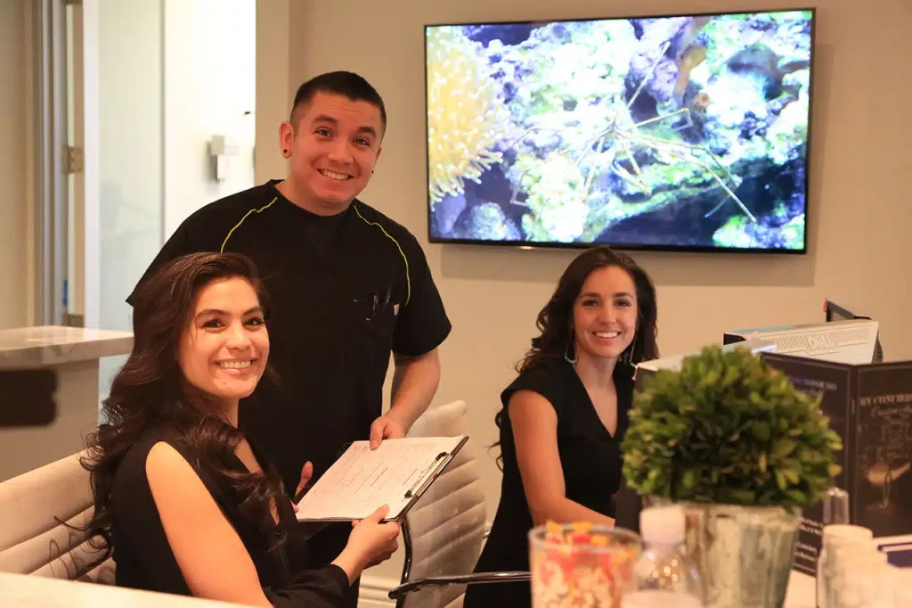 Three people, two women and one man, are smiling at a reception desk with paperwork; an aquarium is displayed on a TV screen in the background.