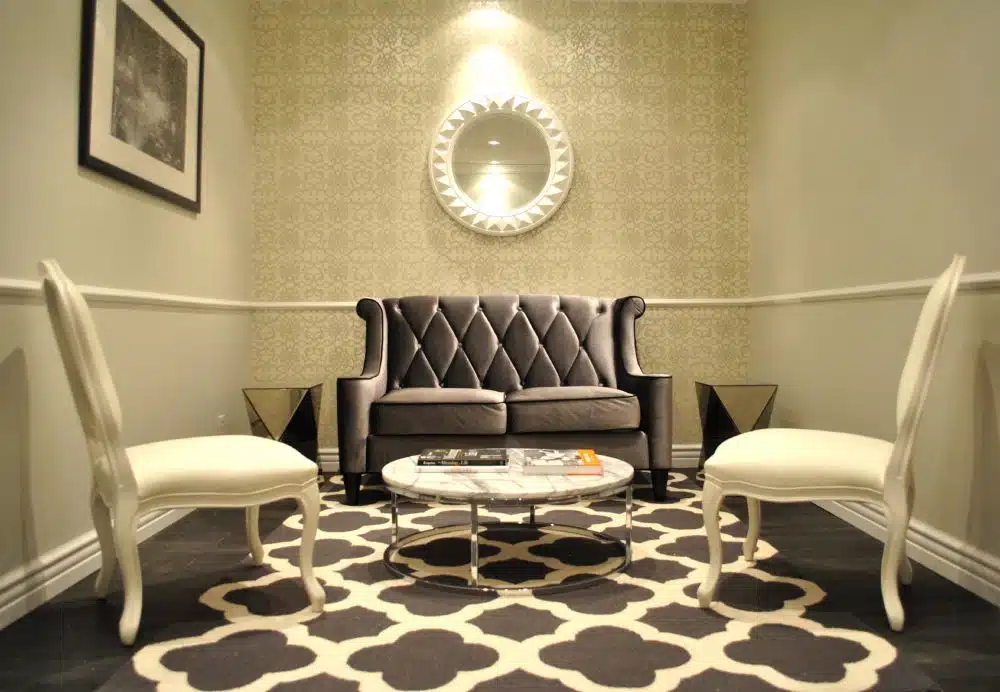 A small sitting area with a gray tufted loveseat, two white chairs, a round coffee table, patterned rug, and a decorative mirror on the wall.
