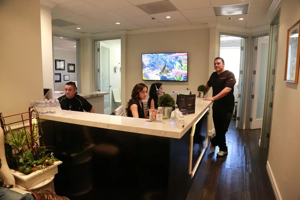 Reception area of an office with three people seated at a desk and one person standing nearby; a TV screen and decor are visible in the background.