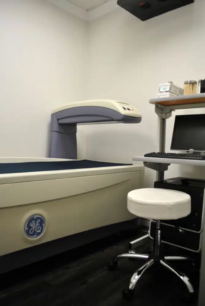 A medical imaging machine next to a computer workstation and stool in a clinical examination room.