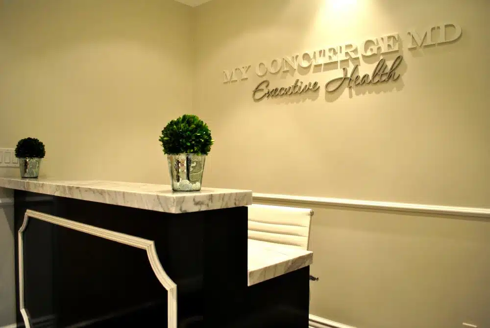 Reception area with a black and white marble desk, two potted plants, and a wall sign reading "MY CONCIERGE MD Executive Health.