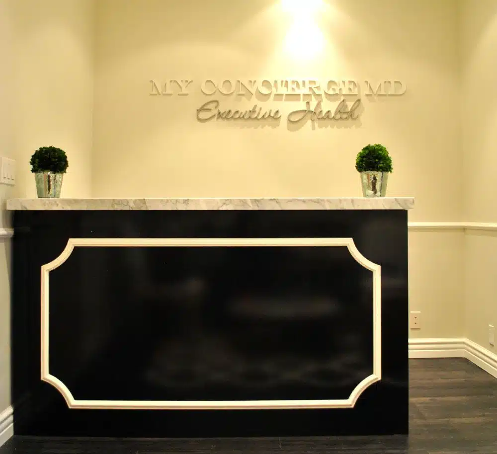 Reception desk with a black front panel, two small potted plants, and a wall sign reading "MY CONCIERGE MD Executive Health" in a well-lit office.