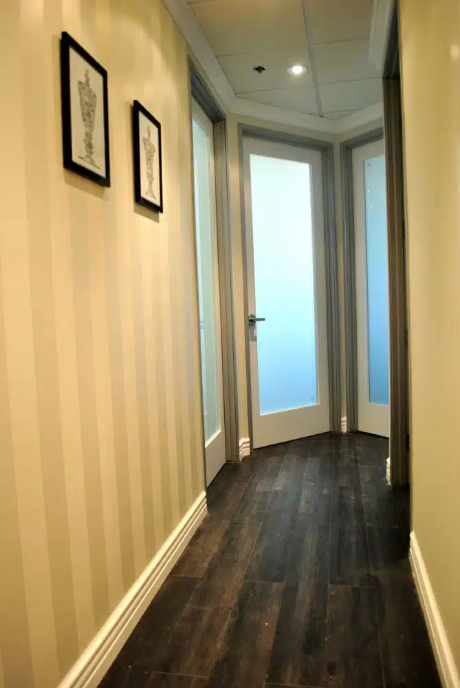 A narrow hallway with striped beige walls, dark wood flooring, two framed artworks, and three frosted glass doors at the end.