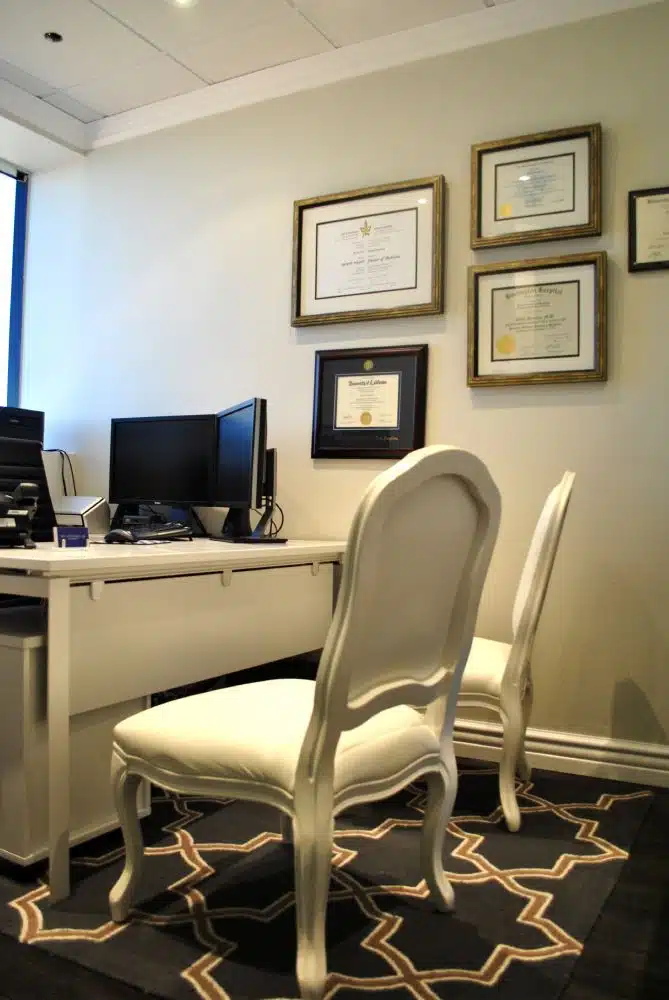 A small office with two white chairs, a desk with a computer, and framed certificates hanging on the wall.
