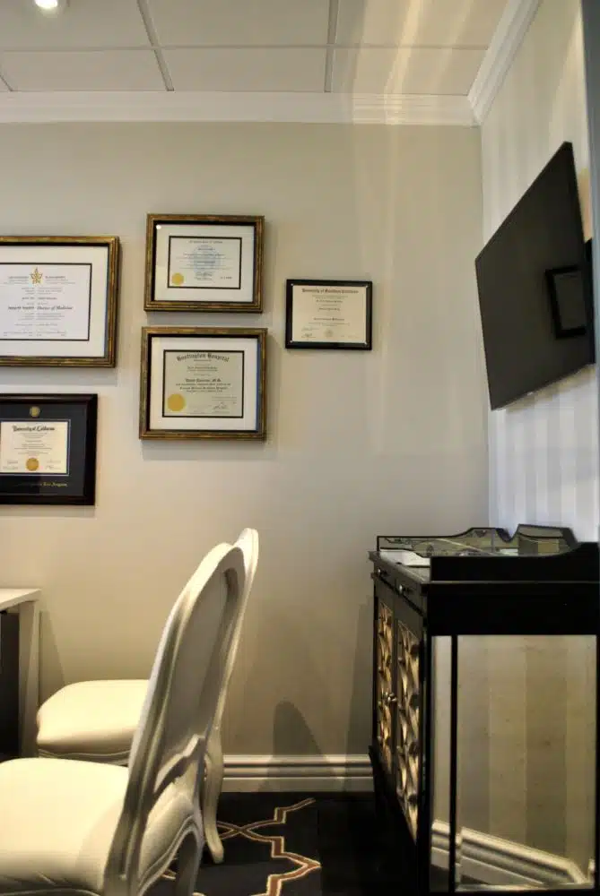 A small office space with two white chairs, framed certificates on the wall, a flat-screen TV, and a mirrored cabinet.
