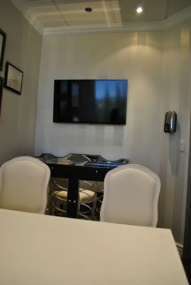 A small office or meeting room with two white chairs, a white table, a black console table, and a flat-screen TV mounted on the wall.