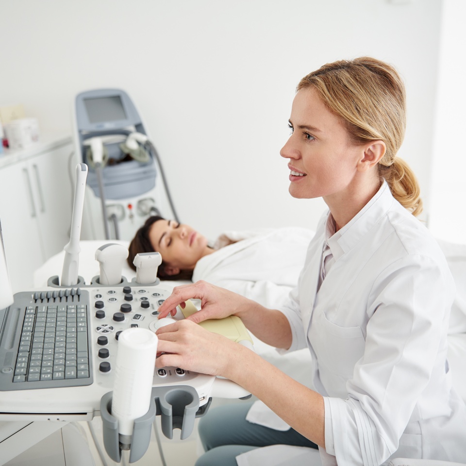 Side view portrait of smiling physician in white lab coat looking at monitor of ultrasound machine. Brunette woman with closed eyes lying on daybed on blurred background