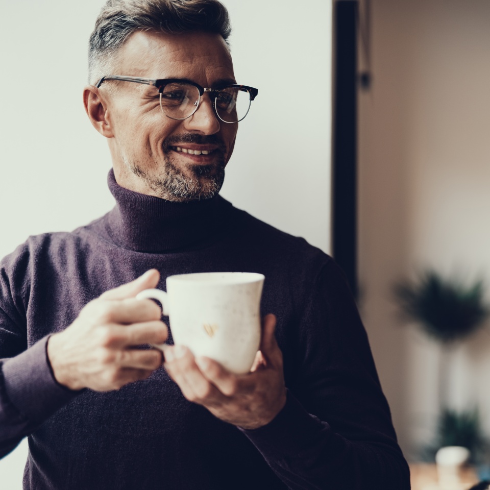 Adult man in casual wear resting in cafe. He standing in with mug of tea against blurred background and smiling