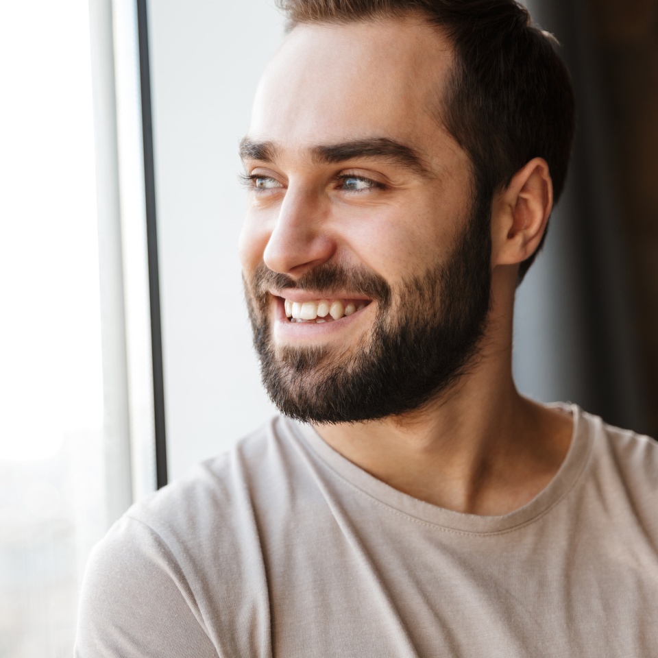 Handsome smiling young bearded man standing at the window at home, looking away