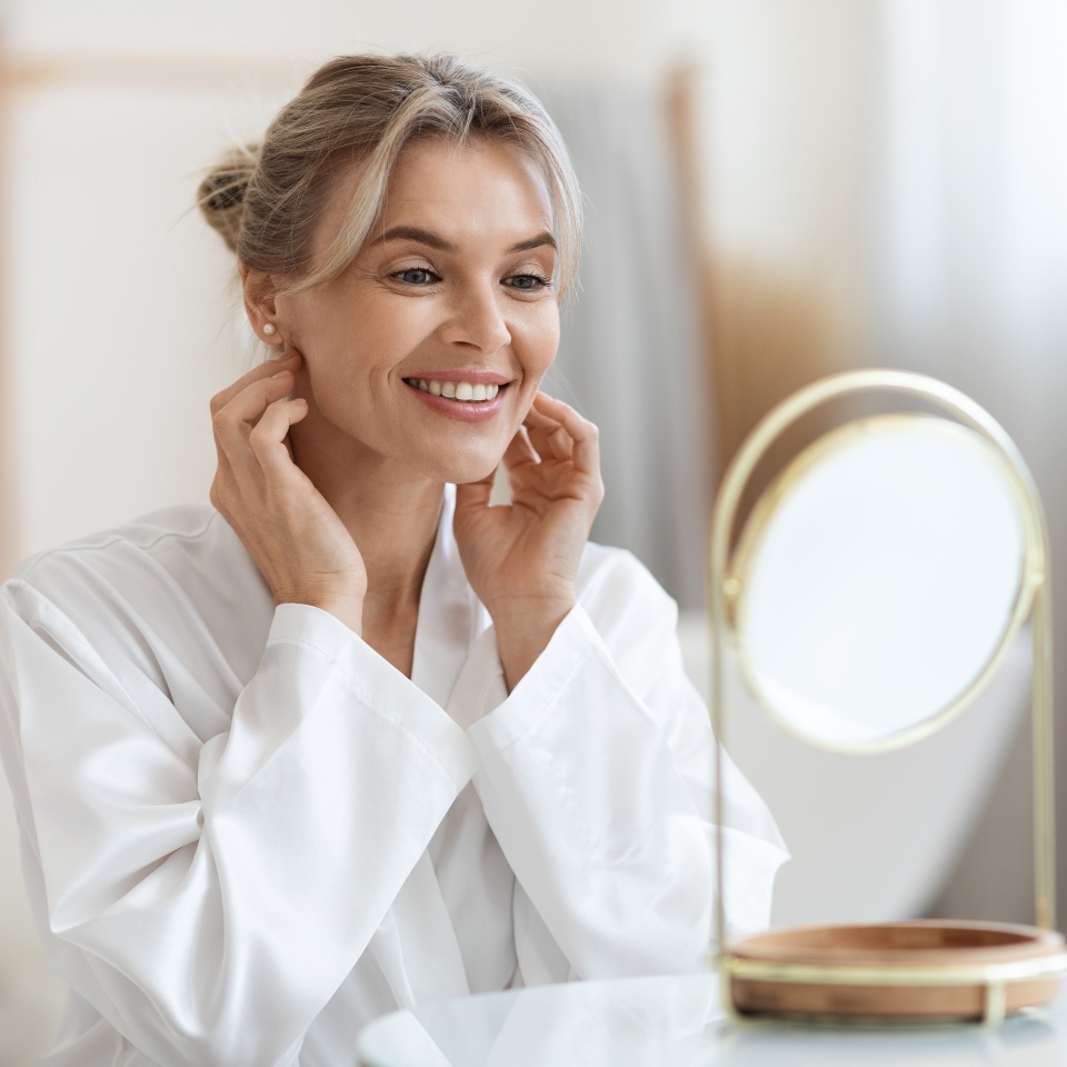Cheerful beautiful blonde middle aged lady in silky bathrobe using hydrating face cream, looking at mirror and smiling, enjoying face care routine, bathroom interior, closeup shot, copy space