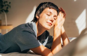 A woman lies on a bed in bright sunlight from the window.