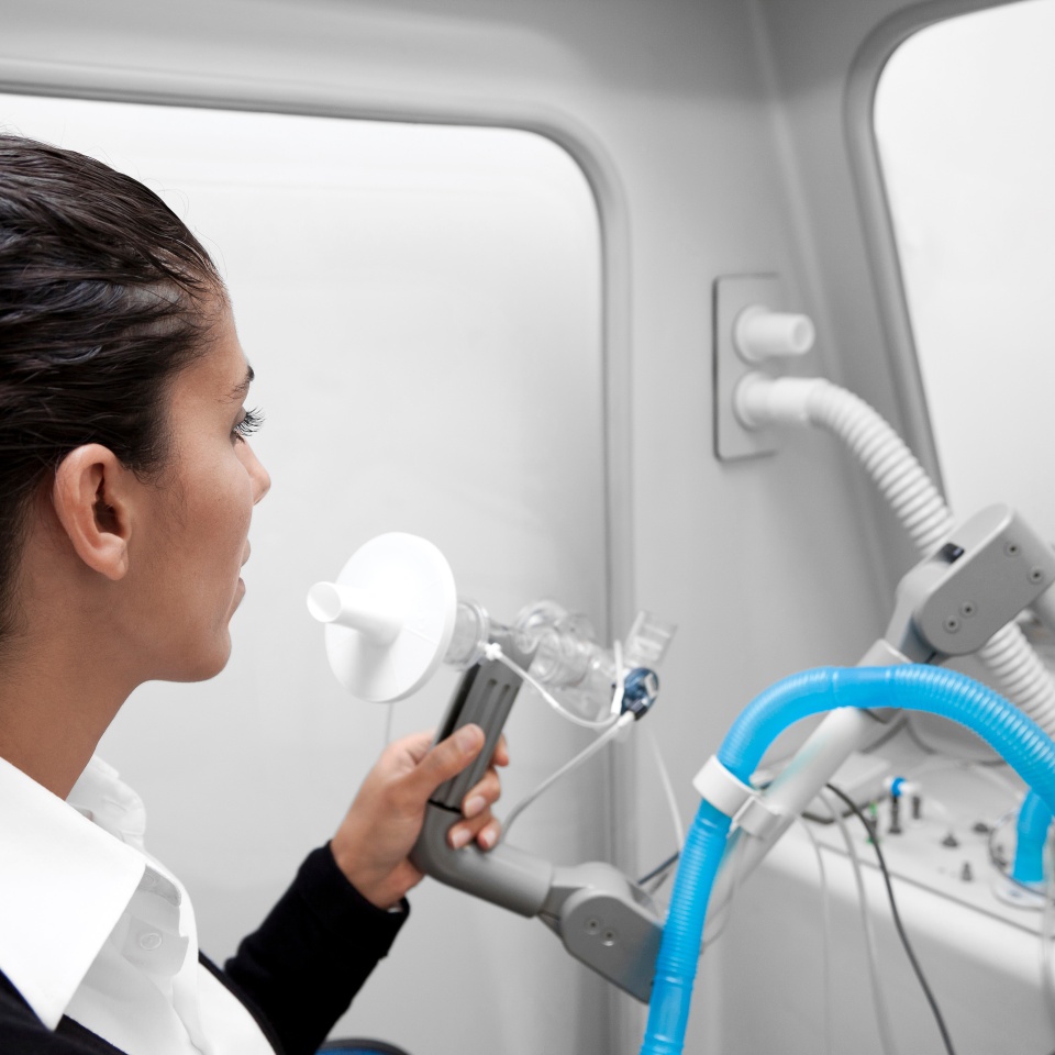 A person uses a medical device, holding a mouthpiece connected to tubes, possibly undergoing a respiratory or lung function test in a clinical setting.