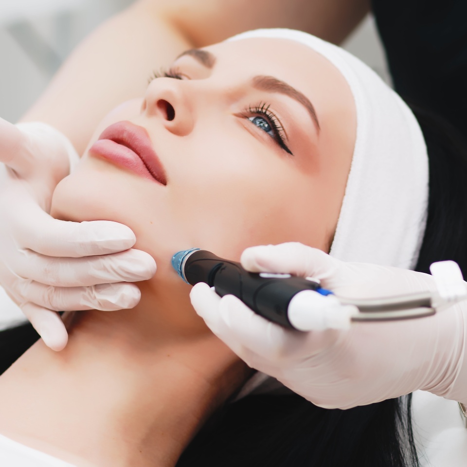 A woman receiving a facial treatment with a handheld device, while wearing a white headband. A professional wearing gloves holds her chin and operates the device on her face.