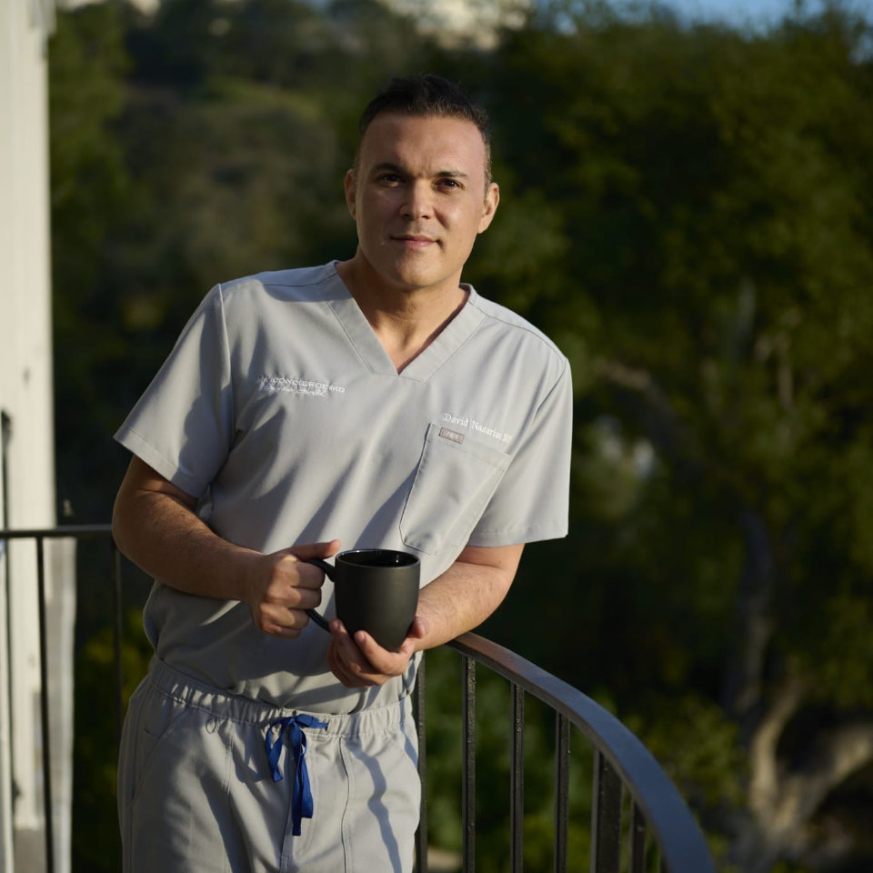 A man in light gray scrubs stands on a balcony holding a black mug, with trees and greenery in the background.