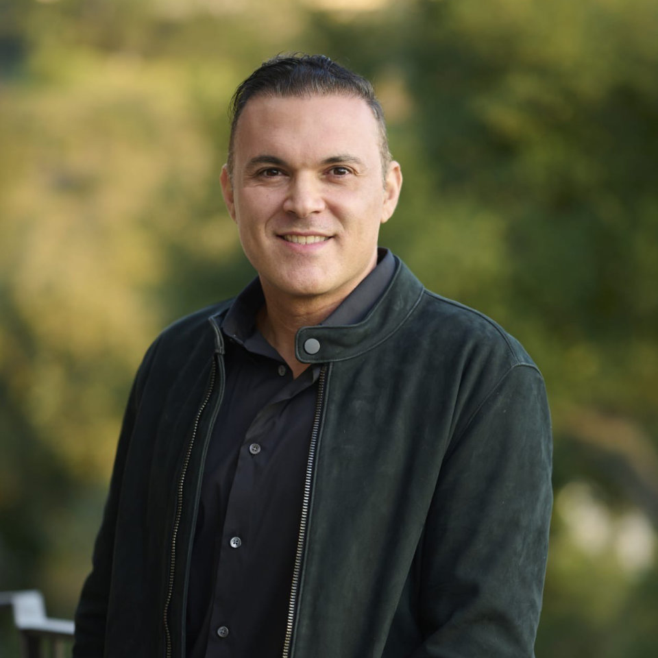A man wearing a dark jacket and shirt stands outdoors, smiling at the camera, with blurred green trees in the background.