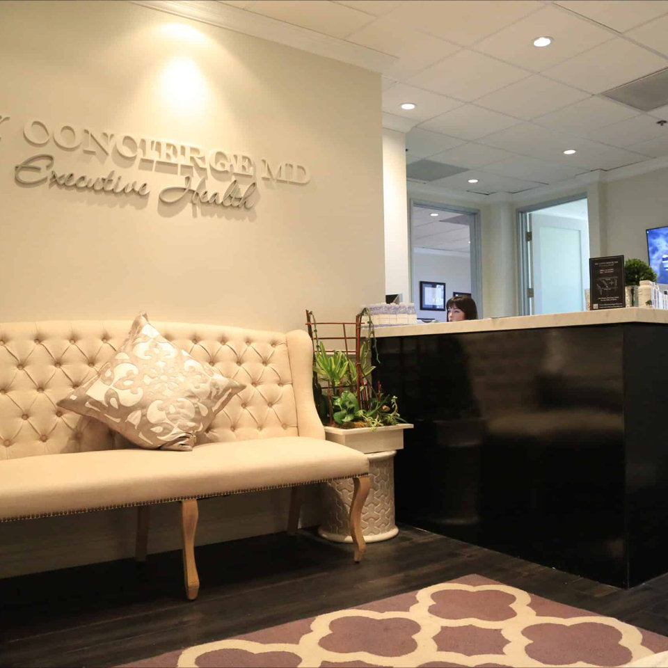 Reception area with a beige tufted sofa, decorative pillow, potted plant, and a black counter; office signage on the wall and a staff member visible behind the counter.
